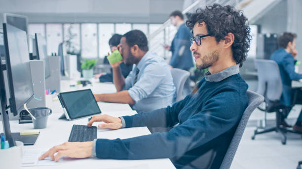 istock Young Handsome Manager with Curly Hair Works on a Desktop Computer and Compares Data with a Software. Diverse and Motivated Business People Work on Computers in Modern Open Office. 1267134390