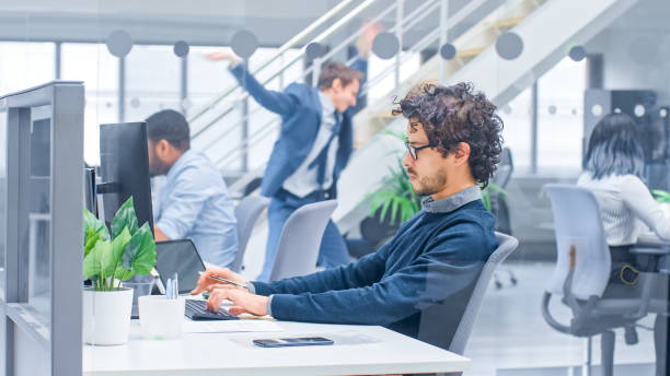 istock Young Handsome Manager with Curly Hair Works on a Desktop Computer and Compares Data with a Software. Diverse and Motivated Business People Work on Computers in Modern Open Office. 1267134444
