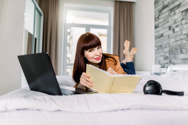 istock Beautiful young smiling woman lying down the bed with legs raised slightly, using laptop and making notes in her notepad. Working and studying from home, stay at home concept 1267137816