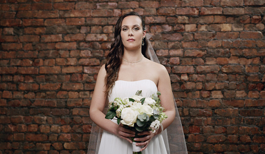 Shot of a beautiful young woman holding a bouquet while standing against a brick wall on her wedding day