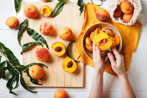 Female hands cut fresh delicious ripe peaches into a plate. Peaches on a cutting board with peach leaves. Flat lay on a white table with an orange towel