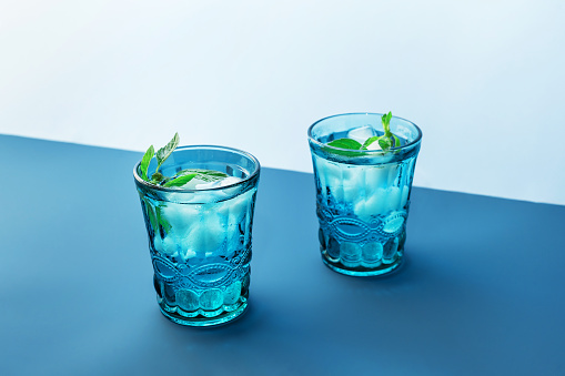 Two beautiful vintage blue glasses with cold drink and ice cubes, with fresh green mint leaves on deep blue and light blue background. Backdrop for your design.