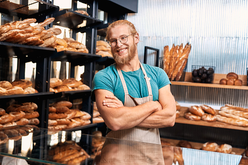Young bearded man wearing eyeglasses and apron assistant standing at bakery shop small business crossed arms looking aside smiling cheerful