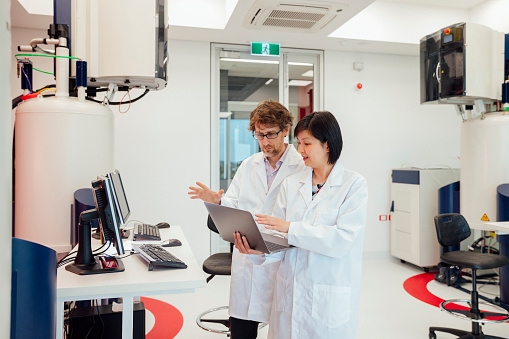 Two scientists standing in a laboratory. There is a lot of large equipment surrounding them. They are wearing lab coats. A Nuclear Magnetic Resonance (NMR) spectroscopy is next to them, this analyses mixture containing known compounds for diseases such as Alzheimer's, diabetes, and heart disease.