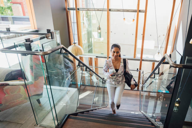 A tilted angle shot of a woman smiling in casual business wear walking up the stairs of a boutique hotel. She has a handbag over her shoulder and her phone in hand.