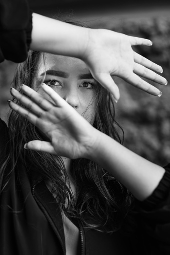 Girl covers her face with her hands and looks through hands. White and black portrait