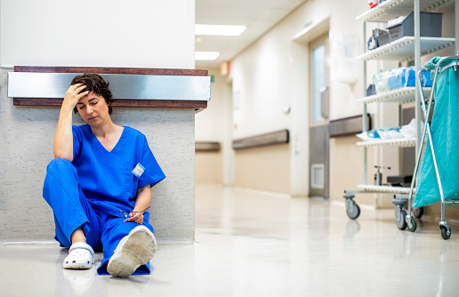 Tired young female doctor in scrubs sitting on the floor of a hospital corridor after a long shift