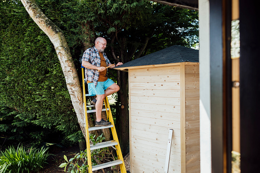 A shot of a senior man on step ladders fixing felt to the roof of a summer house in his garden.