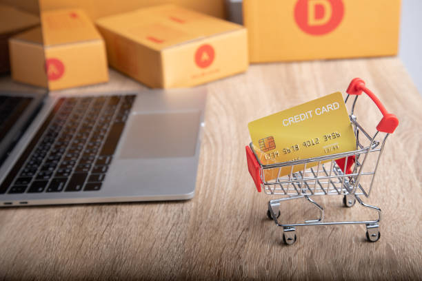 Close-up shot of mock-up credit card in shopping trolley with blurred background of laptop and packaged boxes on wooden table with copy space for ecommerce business concept in the digital technology. toma de primer plano de tarjeta de crédito de maqueta en carro de compras con fondo borroso de computadora portátil y cajas empaquetadas en la mesa de madera con espacio de copia para el concepto de negocio de comercio electrónico en la tecnología digi