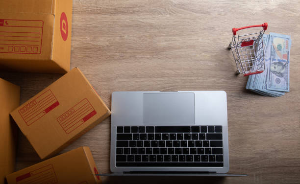 istock Top-view shot for ecommerce business concept in the digital world shows computer laptops, shopping trolley, packaged boxes and many fake 100 US dollar banknotes on wooden table with copy space. 1267162431