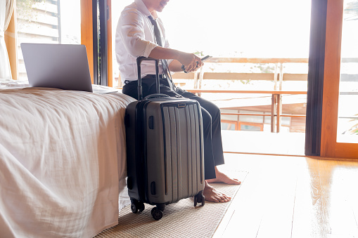 Businessman holding cellphone while sitting on bed with suitcase in hotel room during business trip