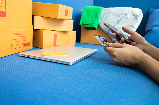 Close-up shot of the teenage shopper is laying down on the sofa, and shopping online via smartphone with credit card for digital technology world and seeing a lot of boxes beside her with the clothes.