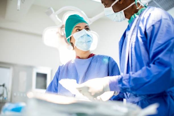 Two diverse doctors prepping for an operation in a hospital Two diverse doctors in scrubs and protective face masks talking together while preparing for a medical procedure in an operating room critical care photos stock pictures, royalty-free photos & images