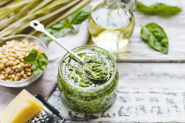 Green fettuccine and Pesto sauce in vintage spoon on glass jar of pesto sauce with ingredients on rustic white wooden table. Traditional Italian pesto recipe for making fettuccine, pasta, bruschetta