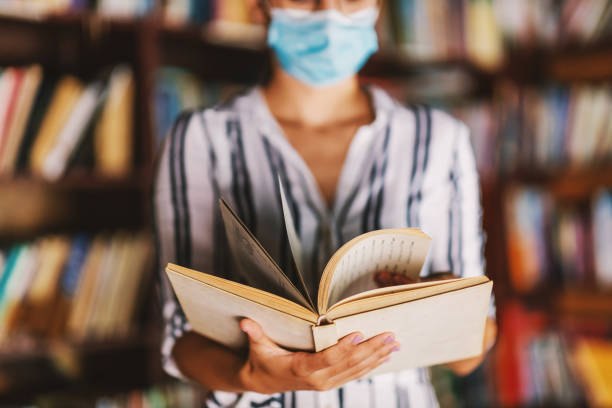 junge college-mädchen mit gesichtsmaske auf stehen in der bibliothek und halten ein buch während der corona-pandemie. - professor librarian university library stock-fotos und bilder