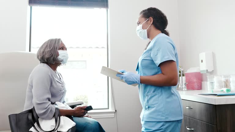 An African American senior woman touches her head and makes other gestures as she discusses her symptoms with a female doctor or nurse. The doctor attentively listens and responds as the patient discusses her symptoms.