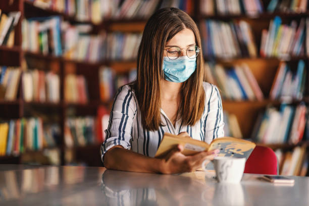 junge attraktive college-mädchen mit gesichtsmaske sitzen in der bibliothek und studieren während der corona-pandemie. - professor librarian university library stock-fotos und bilder