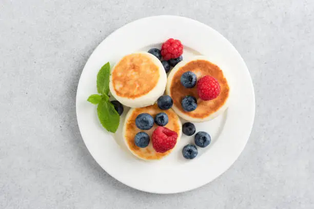 Syrniki with berries on white plate isolated on grey concrete background, top view. Cottage cheese fritters