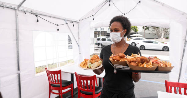 Young Waitress Serving Food to Customers in Outdoor Tent Wearing a Mask A young black woman serves food to customers sitting inside a tent. She is wearing a mask outdoor dining photos stock pictures, royalty-free photos & images