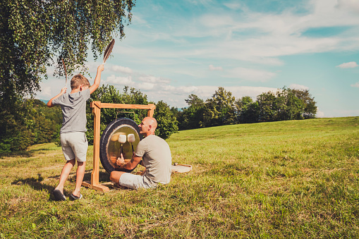Father and son playing therapeutic instruments, gong and shamanic drum in nature.