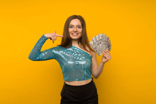 joyful girl won the lottery and pointing on fan of us dollars in her hands on a yellow background - smiling casino human hand beautiful imagens e fotografias de stock