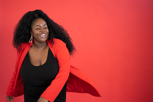 Studio shot of cheerful young black woman with long curly hair wearing unstructured jacket over black scooped neck top and dancing against red background.