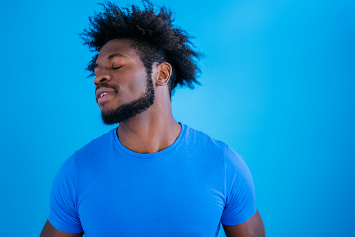 Candid front view of bearded 23 year old black man with half-shaved hairstyle wearing royal blue t-shirt and blissful expression against blue background.