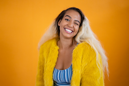 Studio shot of fun early 20s Pacific Islander woman with long two-toned hair in fake fur jacket over striped top smiling at camera against orange background.