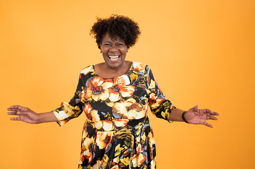 Front view of joyful senior black woman with short curly hair wearing floral print dress and grinning at camera with open arms against orange background.