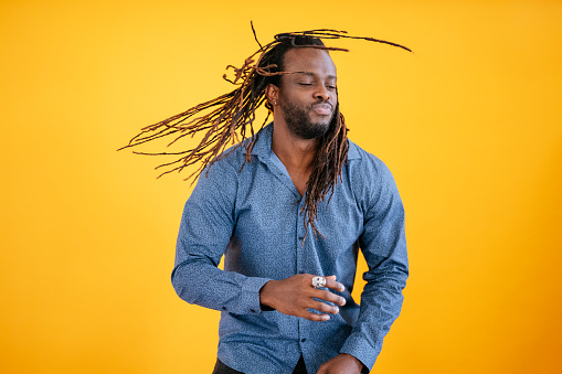 Front view of mid adult black man with beard, mustache, and long dreadlocks in casual open collar shirt moving against orange background.