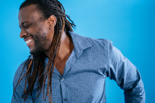 Portrait of smiling early 30s black man with long dreadlocks wearing casual shirt looking away from camera with eyes closed against blue background.
