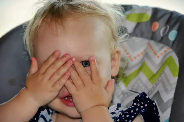 Adorable blonde caucasian baby playing peek-a-boo with one blue eye showing. Hands are covering her face and she is peaking through her fingers.