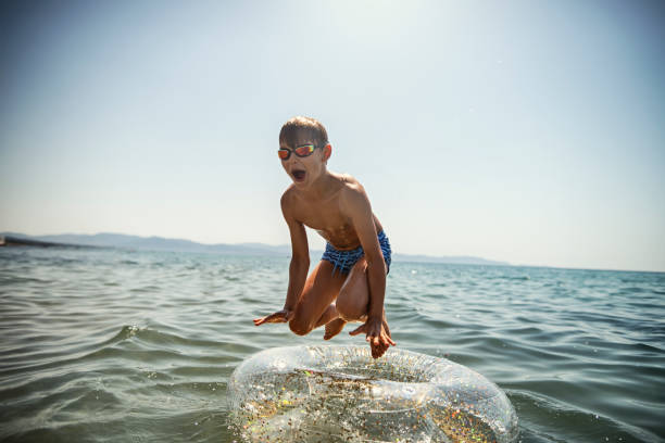 Little boy having fun in the sea. The boy is jumping from  an inflatable ring and falling down on it. Sunny summer day.\nNikon D850