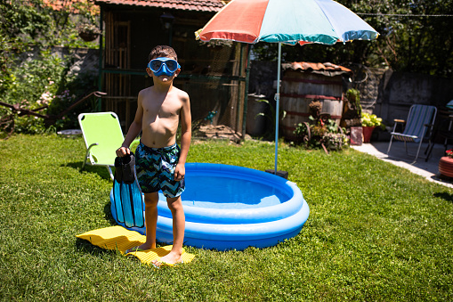 A young boy with diving goggles is standing in front of an inflatable pool holding a pair of diving fins in his hand