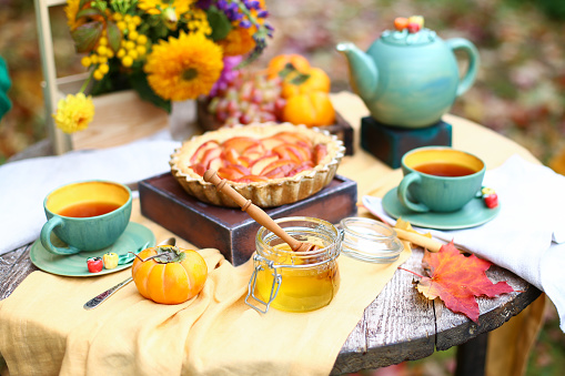 Autumn picnic. Tea party with beautiful kettle, cups at wooden table in garden. Harvest festival. Honey with stick, spoon, apple pie, persimmons, grapes, maple leaf, flowers, yellow linen tablecloth