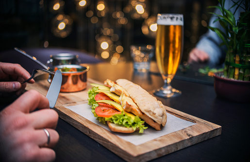 A man in the restaurant has a sandwich served on a wooden tray with salad and meat, on a black background Holding a knife and a fork in his hands with a big beer for a drink.