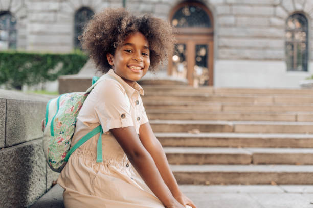istock Cute school girl with a backpack sitting in front of her school 1267218021