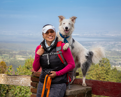 smiling mexican woman with happy white border collie dog standing behind with paws on her shoulder, landscape behind them