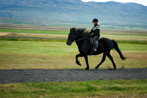 An 11 year old girl  wearing a helmet and traditional woolen sweater  riding a horse on her horse farm, Skagafjordur, Iceland, Europe