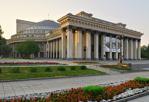 The largest Opera and ballet theater in Russia, the main facade, high columns of the entrance portico, a powerful cornice, a huge dome, the architectural style of Soviet classicism. Green square in the center of the Siberian capital, morning pink light, bright flowers and lawns in summer, empty paved paths in the early morning, without people . Novosibirsk, Siberia, Russia, 2020