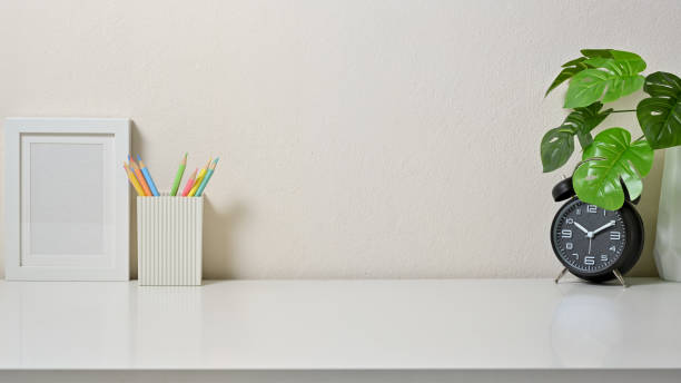 istock Modern workspace with frame picture, coloured pencil, clock and vase on white table in office. Front view. 1267250152