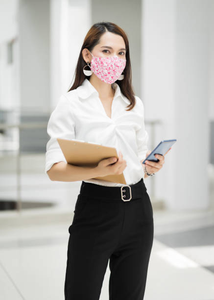 Portrait of a cheerful confident businesswoman in business suit  wears fashion faicial mask to prevent COVID-19, holds document file and cellphone in the business building. Business stock photo stock photo