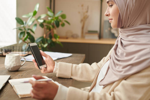современная мусульманская бизнесвумен в платке сидит за офисным столом, проверяя сообщения на своем смартфоне - women middle eastern ethnicity islam hijab стоковые фото и изображения