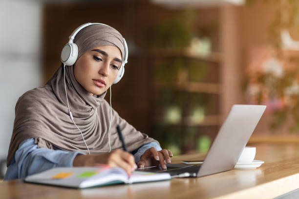 Girl in headscarf having online lesson on laptop at cafe Girl in headscarf having online lesson on laptop at cafe, wearing headset, taking notes, online education concept, copy space religious dress stock pictures, royalty-free photos & images