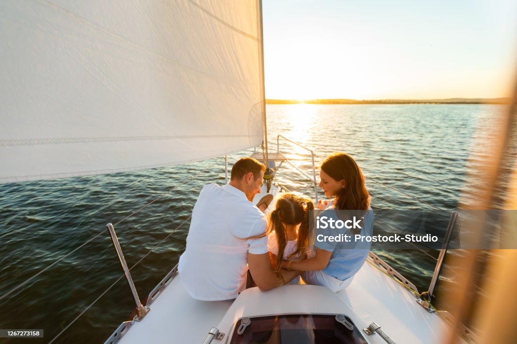 Family Yacht Sailing, Parents And Daughter Sitting On Deck, Back-View Family Yacht Sailing. Parents And Little Daughter Sitting Together On Sailboat Deck Hugging Enjoying Sea Trip. Back View, Free Space Family Stock Photo