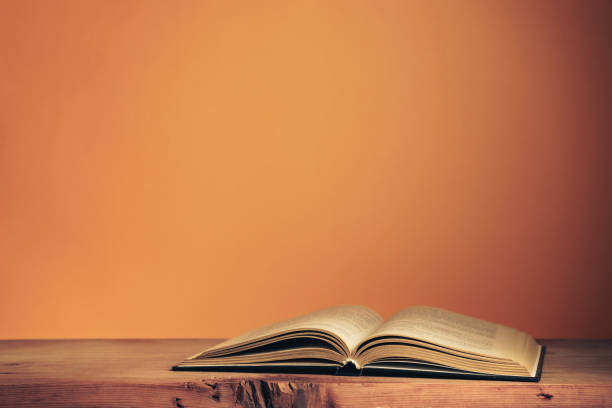 istock Beautiful ancient open old book on a red  wooden table and orange wall background behind. 1267274528