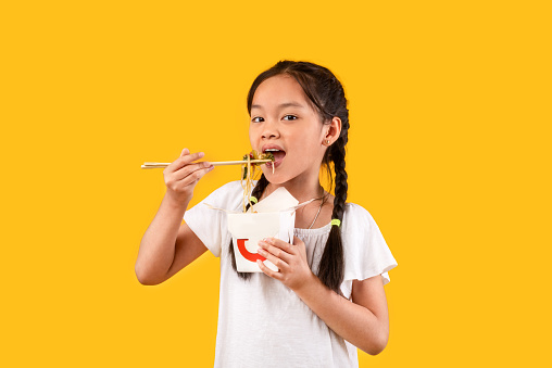 Chinese Kid Girl Eating Noodles With Chopsticks Holding Paper Box Standing On Yellow Studio Background. Fastfood, Restaurant Food Concept