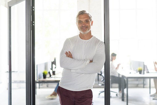Happy businessman standing in the office with coworkers in the background working by the desk