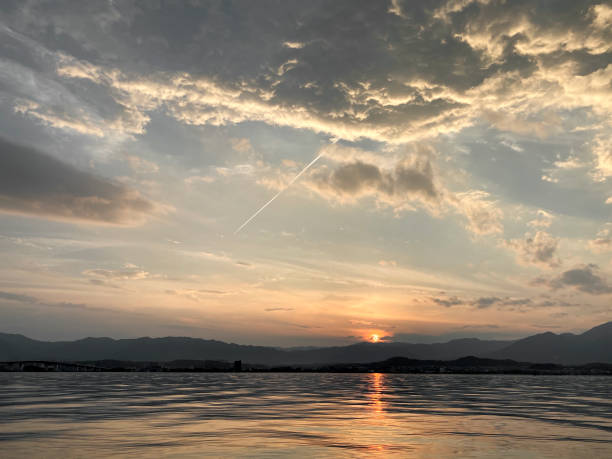 An evening view of the sun, clouds, contrails, and waves on the surface of the lake, which set in the mountains.