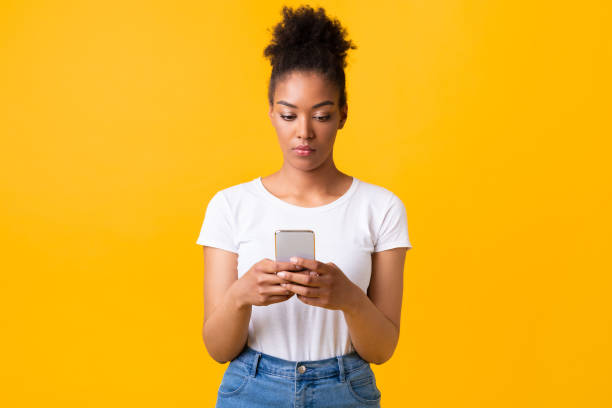 Focused afro girl using cell phone at studio Portrait of serious black lady using her smart phone isolated on orange studio wall serious black teen stock pictures, royalty-free photos & images